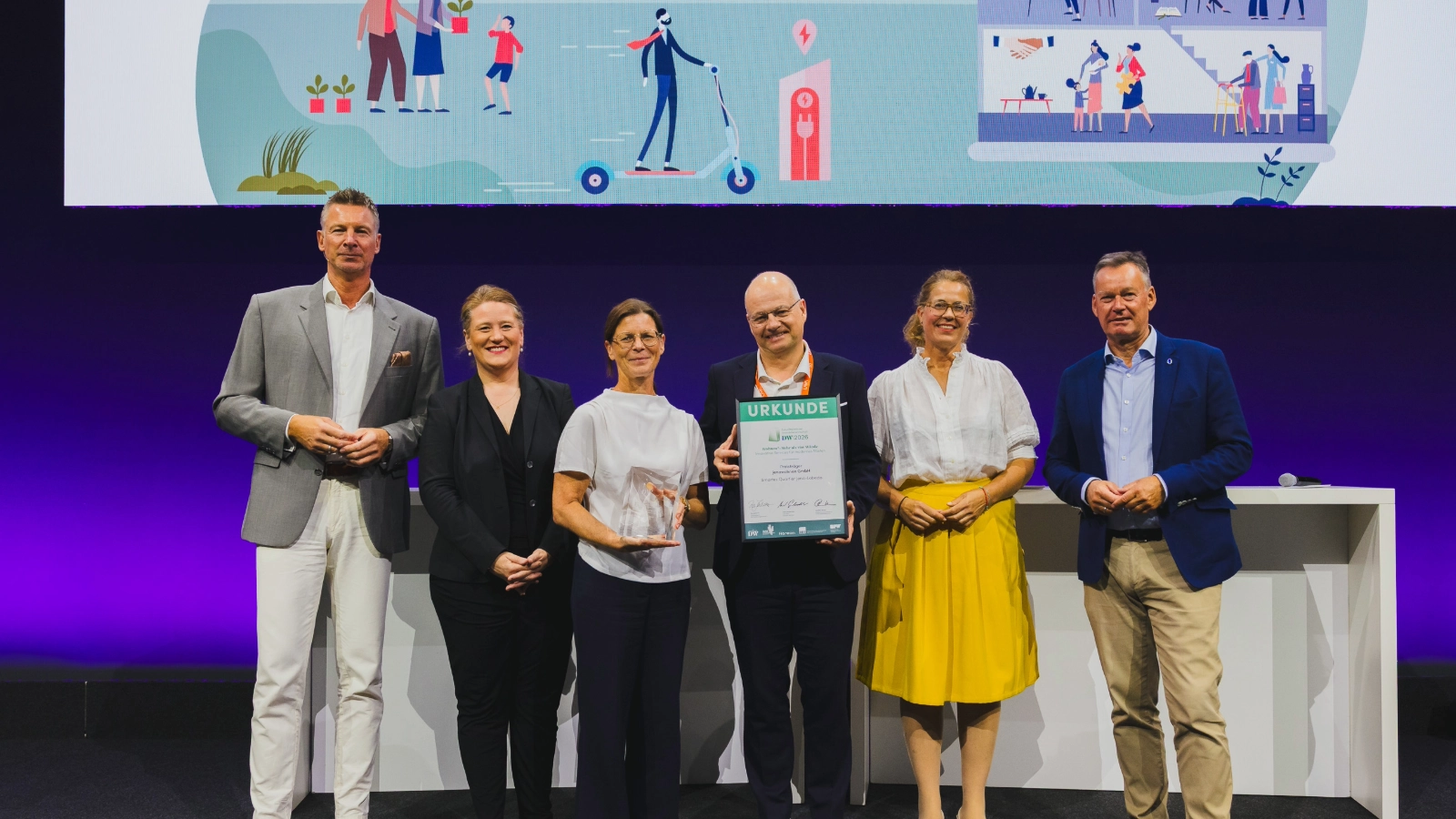 Freuen sich über den DW-Zukunftspreis der Immobilienwirtschaft 2026: Susanne Lippmann, Leiterin Technisches Management jenawohnen, und Tobias Wolfrum, Geschäftsführer jenawohnen und Stadtwerke Jena, gemeinsam mit Vertreterinnen und Vertretern der Jury.