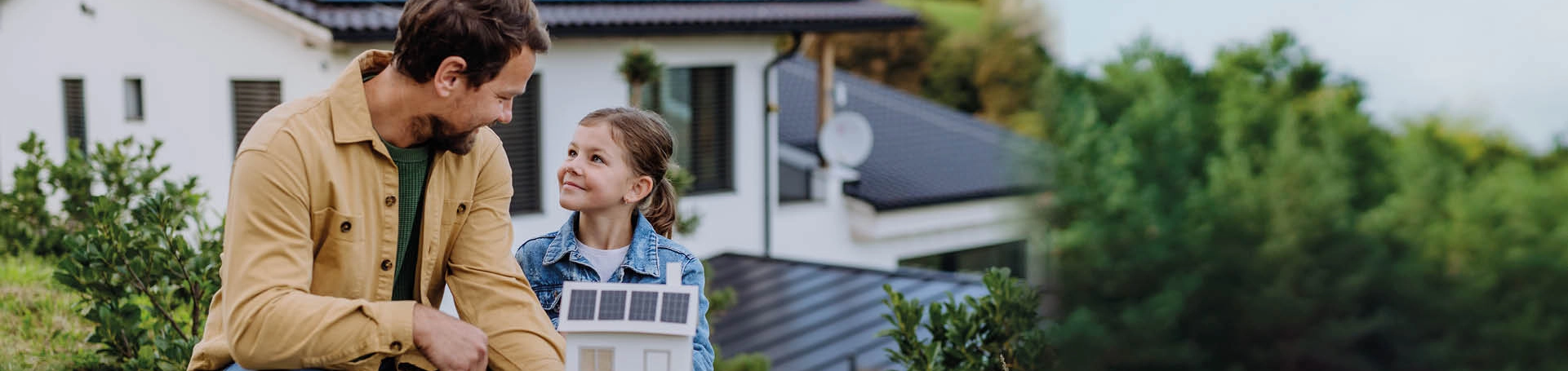 Vater und Tochter vor einem Haus mit Solardach. Er hält ein Miniaturhaus mit Solardach in der Hand.