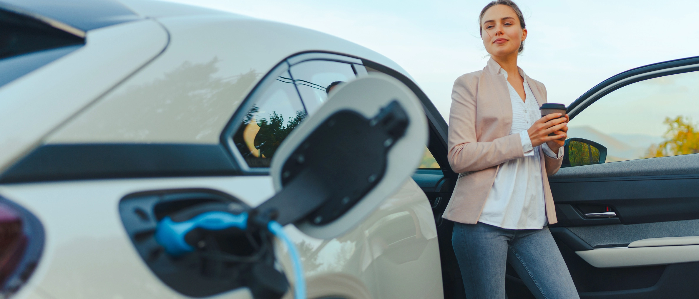 Junge Frau steht in der geöffneten Tür ihres E-Autos und schaut entspannt beim Laden zu.