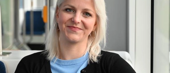 Eine blonde Frau mit hellblauem Pullover sitzt in einer Lichtbahn am Fenster und lächelt in die Kamera.