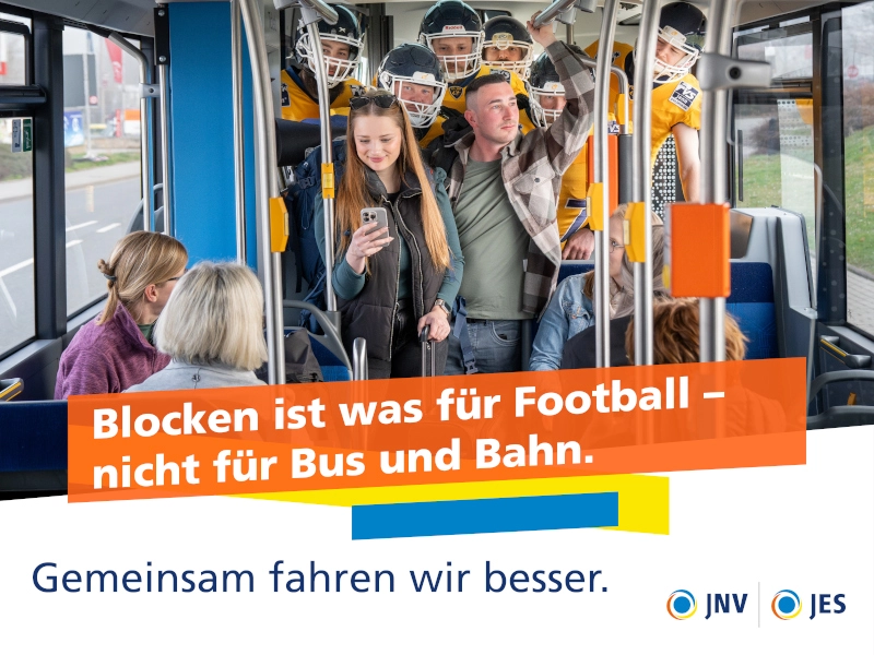 Ein Mann und eine Frau stehen im Mittelgang eines Busses, sie schaut auf ihr Handy, er aus dem Fenster. Dahinter stehen gedrängt Footballspieler, während auf den Sitzplätzen rechts und links die Fahrgäste auf das Pärchen schauen.,